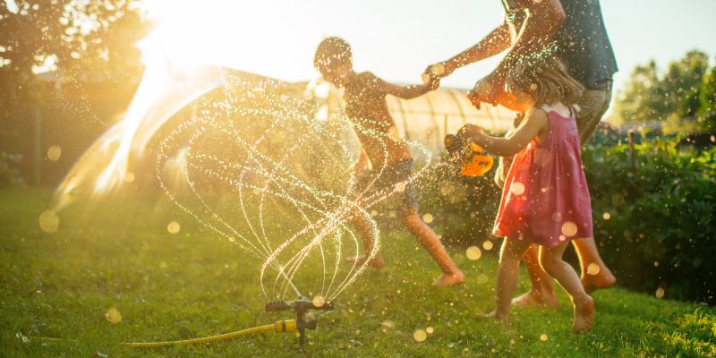 Father with children running through sprinkler at back yard in summer evening in sunset - Mspark summerblog